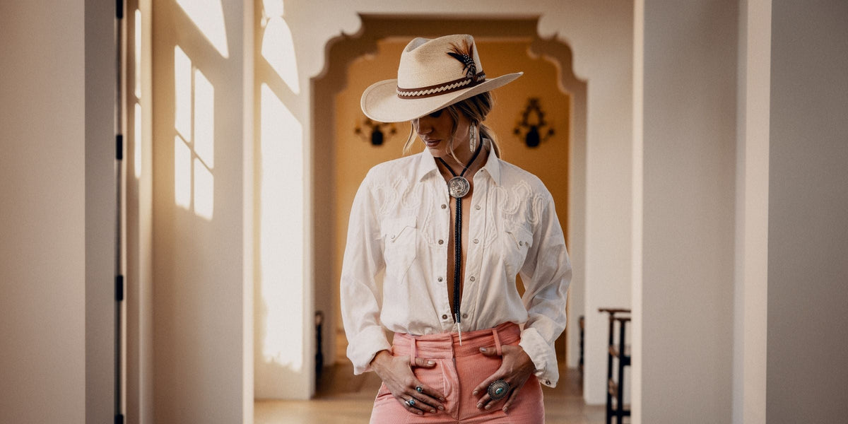 Lainey Wilson wearing the Charlie 1 Horse x Lainey Wilson Road Runner straw cowgirl hat with a white western shirt, bolo tie and pink flared cords
