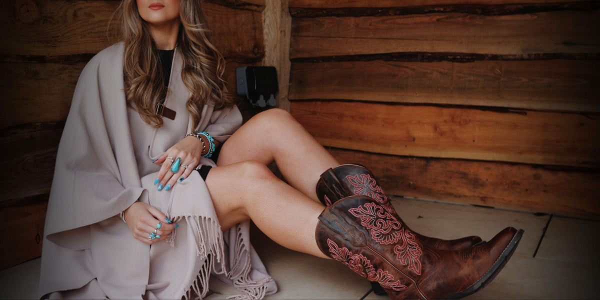 Woman in a cream poncho and turquoise jewellery wearing Ariat Round Up StretchFit cowboy boots with pink floral embroidery, seated against a log cabin wall