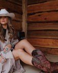 Woman wearing Ariat Round Up StretchFit brown leather cowboy boots with pink floral embroidery, styled with a white hat and beige wrap, seated against a rustic log cabin wall