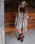Woman wearing Ariat Round Up StretchFit brown cowboy boots with pink floral embroidery, styled with a beige fringed poncho and wide-brimmed hat, standing by a rustic wooden barn wall