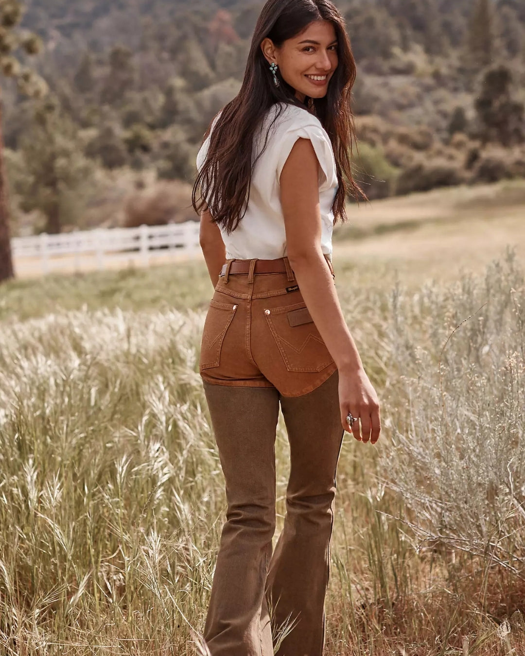 Back view of Wrangler x Lainey Wilson brown chap jeans, showing the tan seat panel and flared legs, styled with a white tee outdoors