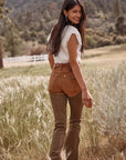 Back view of Wrangler x Lainey Wilson brown chap jeans, showing the tan seat panel and flared legs, styled with a white tee outdoors
