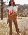 Woman in a wild grass meadow wearing Wrangler x Lainey Wilson brown chap jeans with a white horse graphic tee and western belt