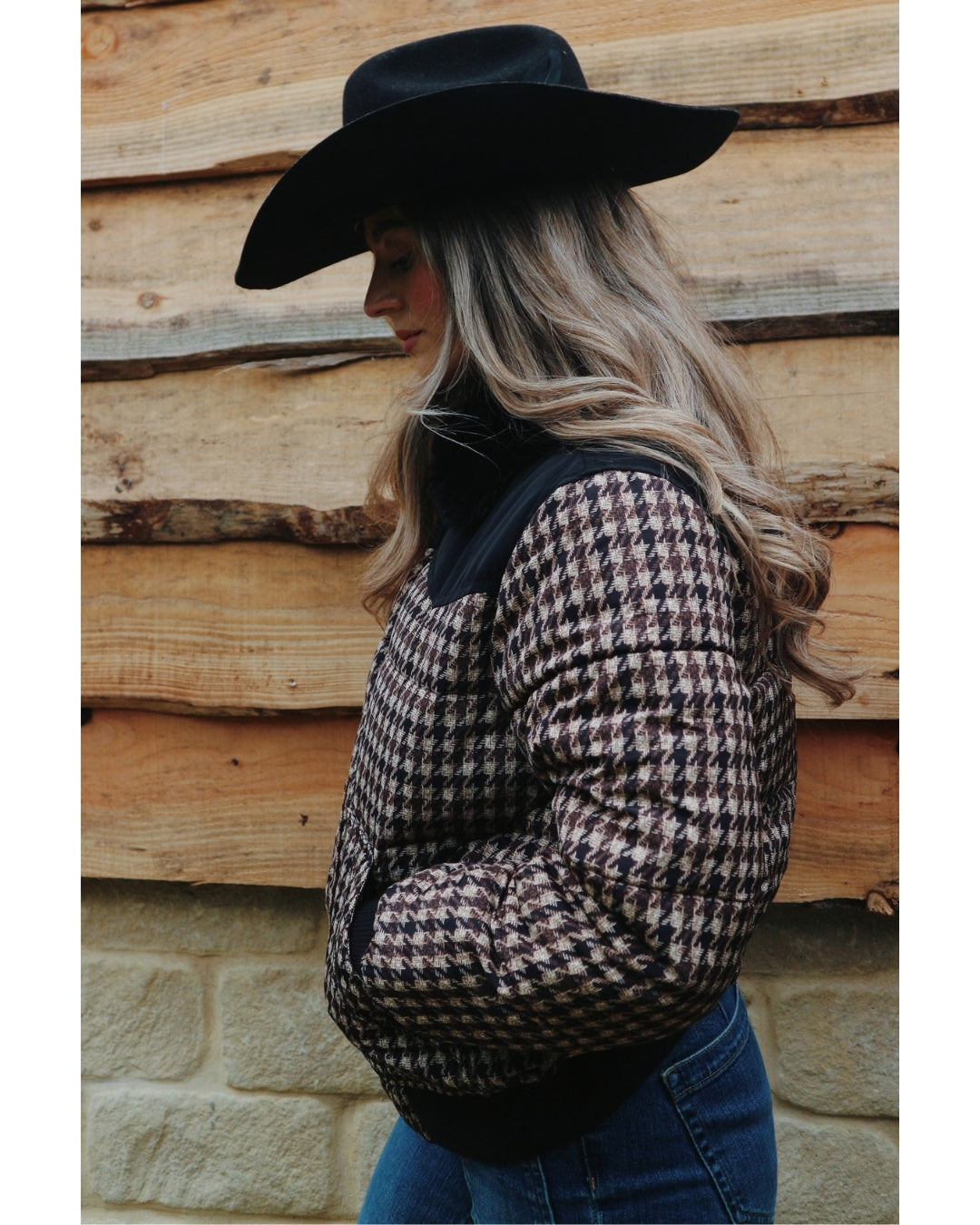 Side view of the Wrangler Western houndstooth puffer jacket styled with a black cowboy hat and blue jeans against a timber wall