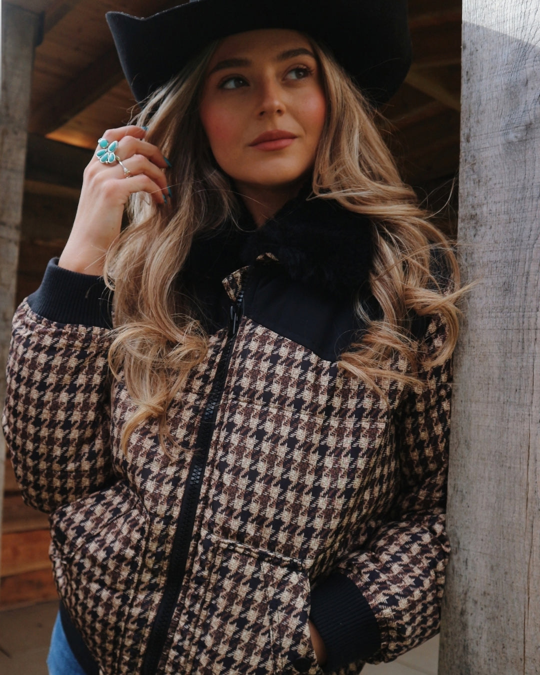 Woman in a black cowboy hat wearing the Wrangler Western houndstooth puffer jacket with black faux fur collar, leaning on a barn post